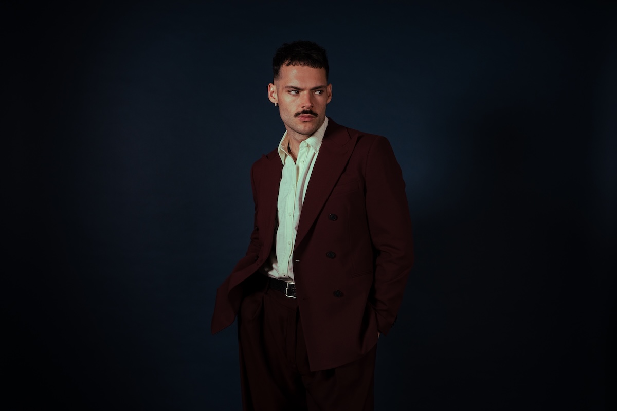 Musician James Marriott wearing a maroon suit and white collared shirt looking to the side against a dark backdrop.